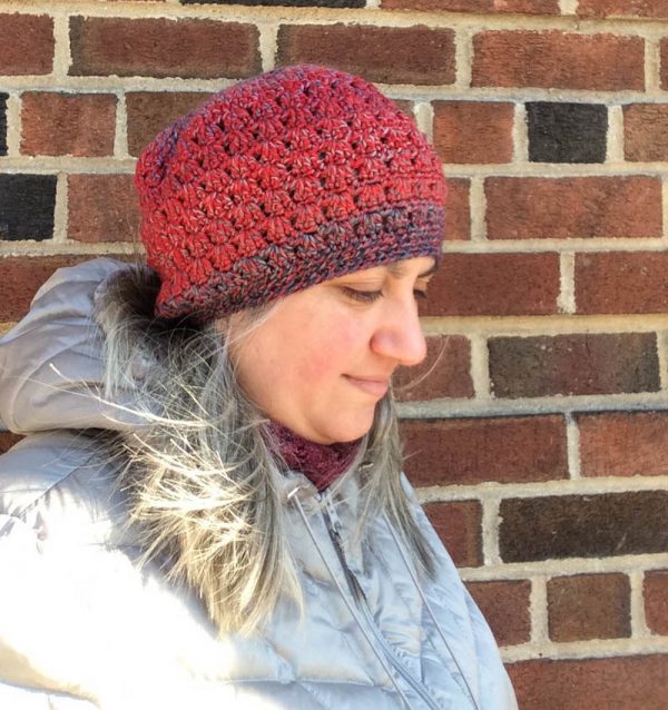 Woman looking down while wearing striped crochet hat and silver coat against a brick wall | Free crochet pattern: Flattened Shell Hat in 4 sizes in Cascade 220 Superwash Wave yarn by Underground Crafter