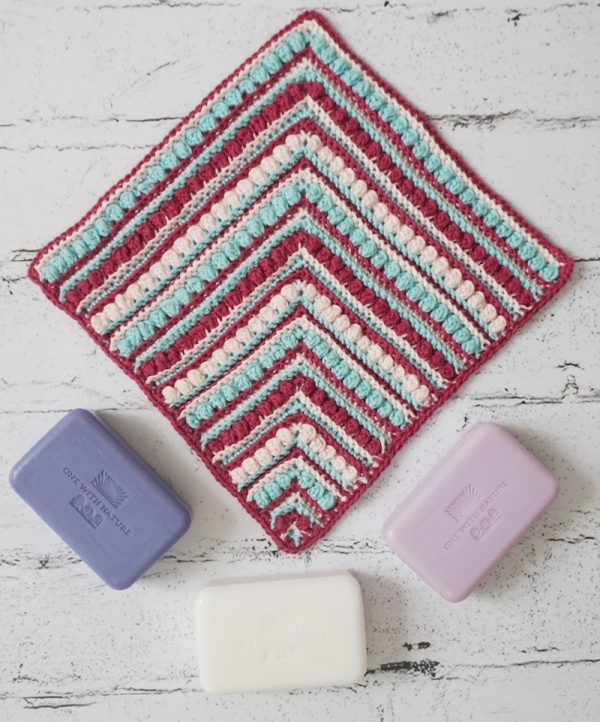 Rosettes and Ridges Washcloth, free crochet pattern in Paintbox Yarns Cotton DK by Underground Crafter | Striped, textured crochet mitered square on faux brick background with soap bars